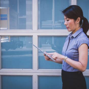 Photograph of an Asian business woman reading something on an ipad, in front of a set of office windows.